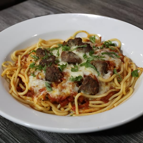 Spaghetti with meatballs, melted cheese, and fresh basil in a white bowl on a wooden table.